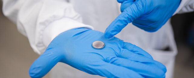 Coin-sized circular silver battery in palm of blue gloved hand, with index finger of the person's other hand pointing to the battery.